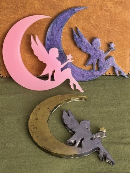 Decorative resin cutouts of faeries on crescent moons in pink, purple, and gold on a green surface.
